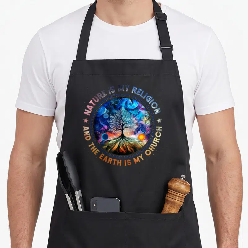 Religion tree apron - Circular cosmic tree of life with sun moon and arched text NATURE IS MY RELIGION AND THE EARTH IS — shown on a black cotton canvas apron against a white background