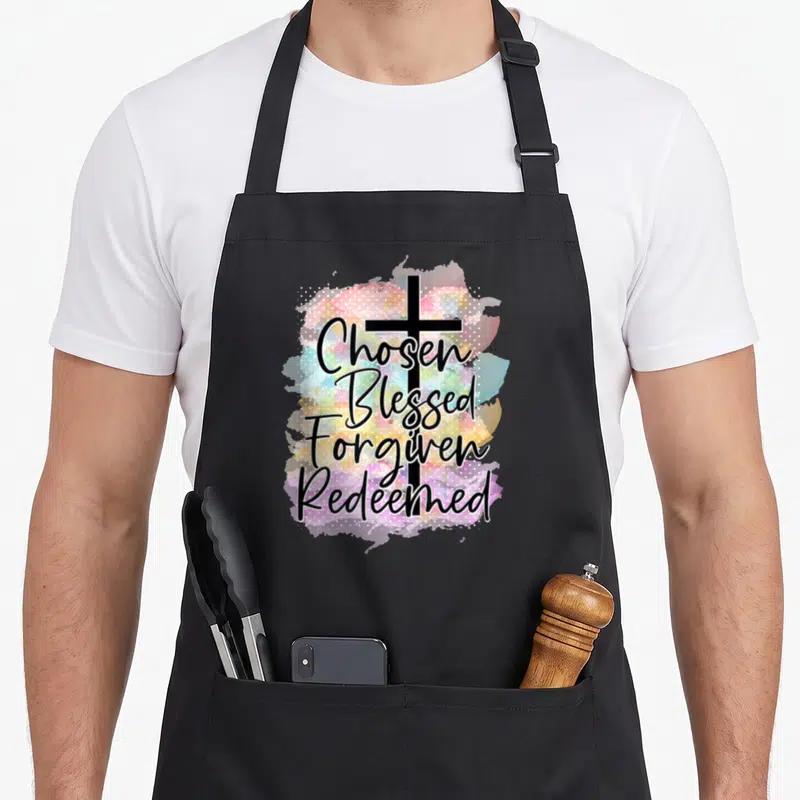 Chosen christian apron - Chosen Blessed Forgiven Redeemed text over black cross with colorful watercolor splash — shown on a black cotton canvas apron against a white background