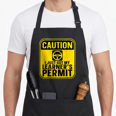Funny learner permit apron - Yellow caution sign with steering wheel and I Just Got My Learner's Permit text — shown on a black cotton canvas apron against a white background