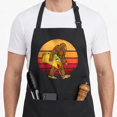 Carrying taco apron - Brown Bigfoot walking right holding giant taco in front of retro striped orange and red sun — shown on a black cotton canvas apron against a white background