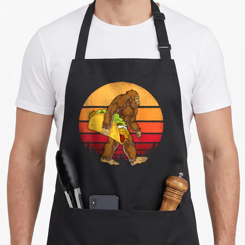 Carrying taco apron - Brown Bigfoot walking right holding giant taco in front of retro striped orange and red sun — shown on a black cotton canvas apron against a white background