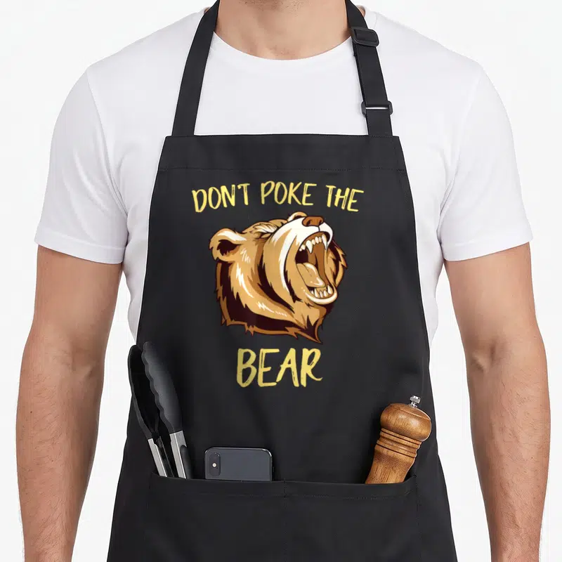 Funny outdoor apron - Cartoon bear head roaring with text Don't Poke The Bear in yellow and brown — shown on a black cotton canvas apron against a white background