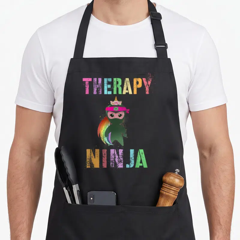 Therapy unicorn apron - Cartoon ninja with rainbow mane and THERAPY NINJA text in pink blue green yellow orange — shown on a black cotton canvas apron against a white background
