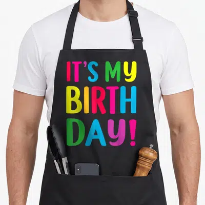 Funny age birthday apron for grilling - IT'S MY BIRTH DAY! text in pink blue green yellow orange stacked vertically — shown on a black cotton canvas apron against a white background