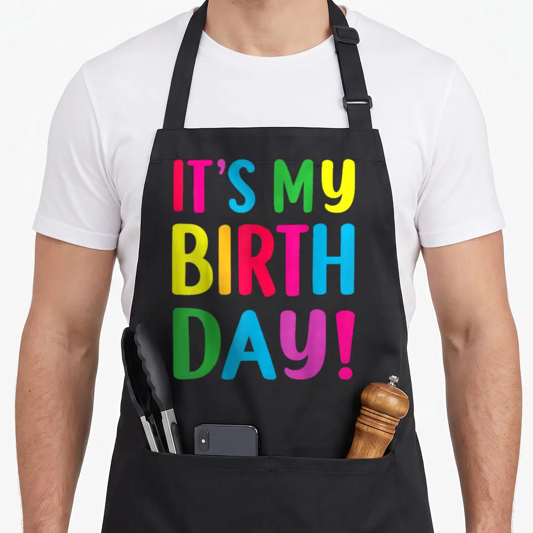 Funny age birthday apron for grilling - IT'S MY BIRTH DAY! text in pink blue green yellow orange stacked vertically — shown on a black cotton canvas apron against a white background