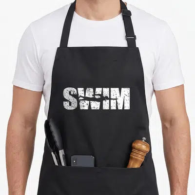 Swimming distressed apron - White distressed SWIM text with grey swimmer silhouette overlapping letters S, W, and I — shown on a black cotton canvas apron against a white background