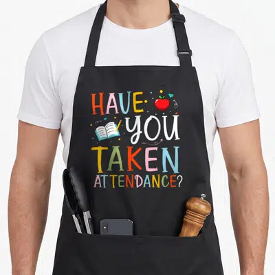 Taken secretary apron - Stacked text HAVE YOU TAKEN ATTENDANCE with red apple, book, pencil, and paper plane icons — shown on a black cotton canvas apron against a white background