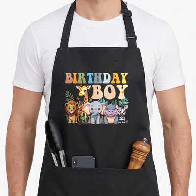 Safari squad birthday apron - Cartoon BIRTHDAY BOY text above lion giraffe elephant hippo zebra snake and foliage — shown on a black cotton canvas apron against a white background
