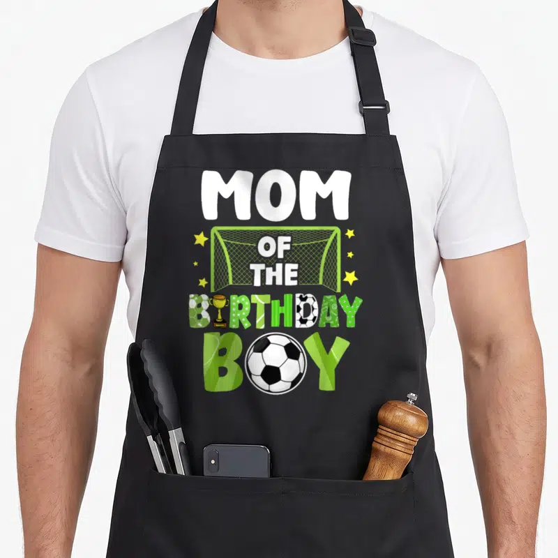 Soccer family aprons - MOM OF THE BIRTHDAY BOY text with soccer goal trophy and ball in green white yellow — shown on a black cotton canvas apron against a white background