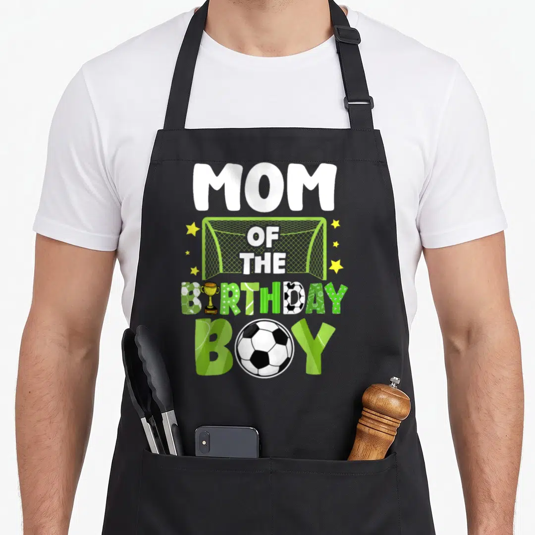 Soccer family aprons - MOM OF THE BIRTHDAY BOY text with soccer goal trophy and ball in green white yellow — shown on a black cotton canvas apron against a white background