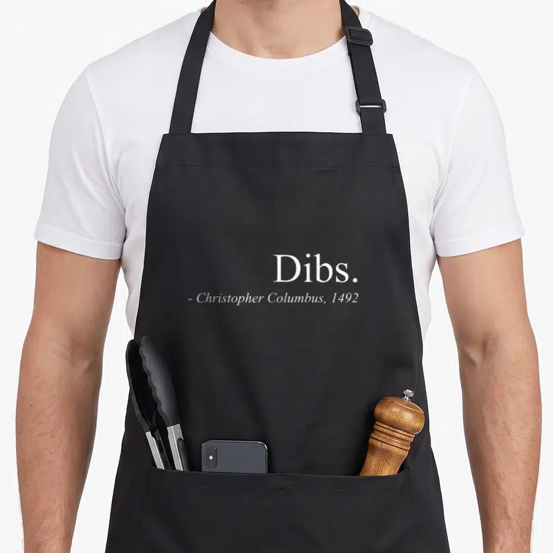 Dib columbus apron - White text Dibs Christopher Columbus 1492 typography on grey background — shown on a black cotton canvas apron against a white background