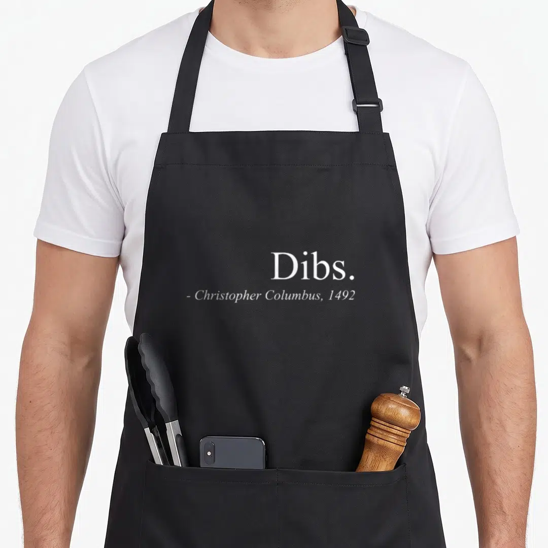 Dib columbus apron - White text Dibs Christopher Columbus 1492 typography on grey background — shown on a black cotton canvas apron against a white background