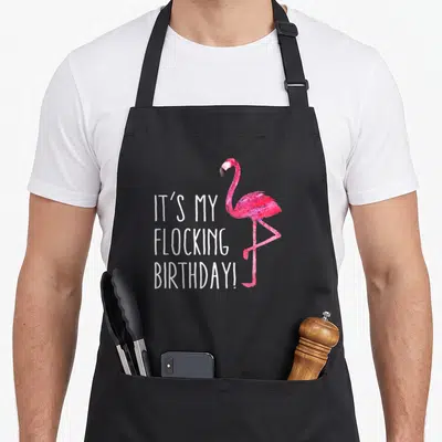 Birthday pink flamingo apron - IT'S MY FLOCKING BIRTHDAY text beside pink watercolor flamingo standing on one leg — shown on a black cotton canvas apron against a white background