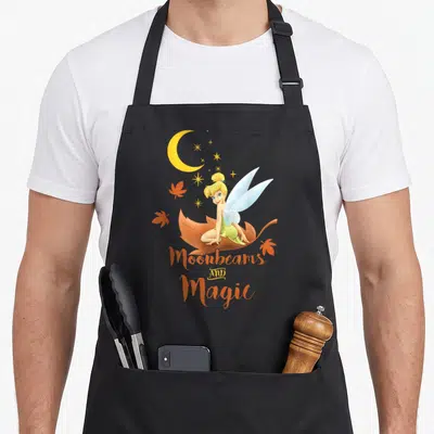 Peter fall apron - Tinker Bell sitting on brown leaf with orange leaves and Moonbeams AND Magic text — shown on a black cotton canvas apron against a white background