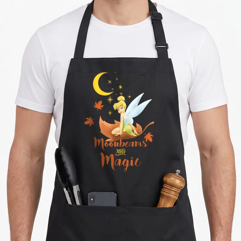 Peter fall apron - Tinker Bell sitting on brown leaf with orange leaves and Moonbeams AND Magic text — shown on a black cotton canvas apron against a white background