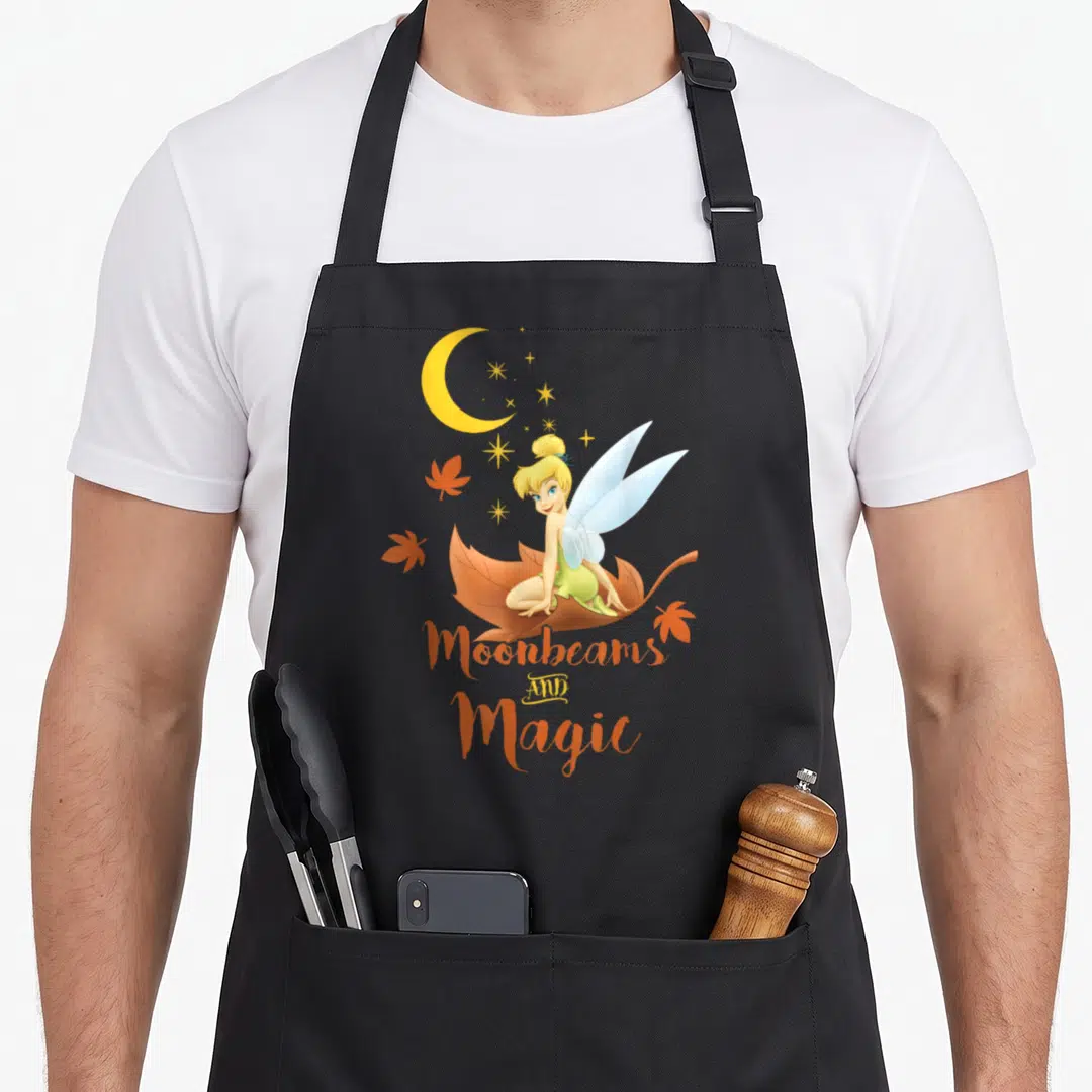Peter fall apron - Tinker Bell sitting on brown leaf with orange leaves and Moonbeams AND Magic text — shown on a black cotton canvas apron against a white background