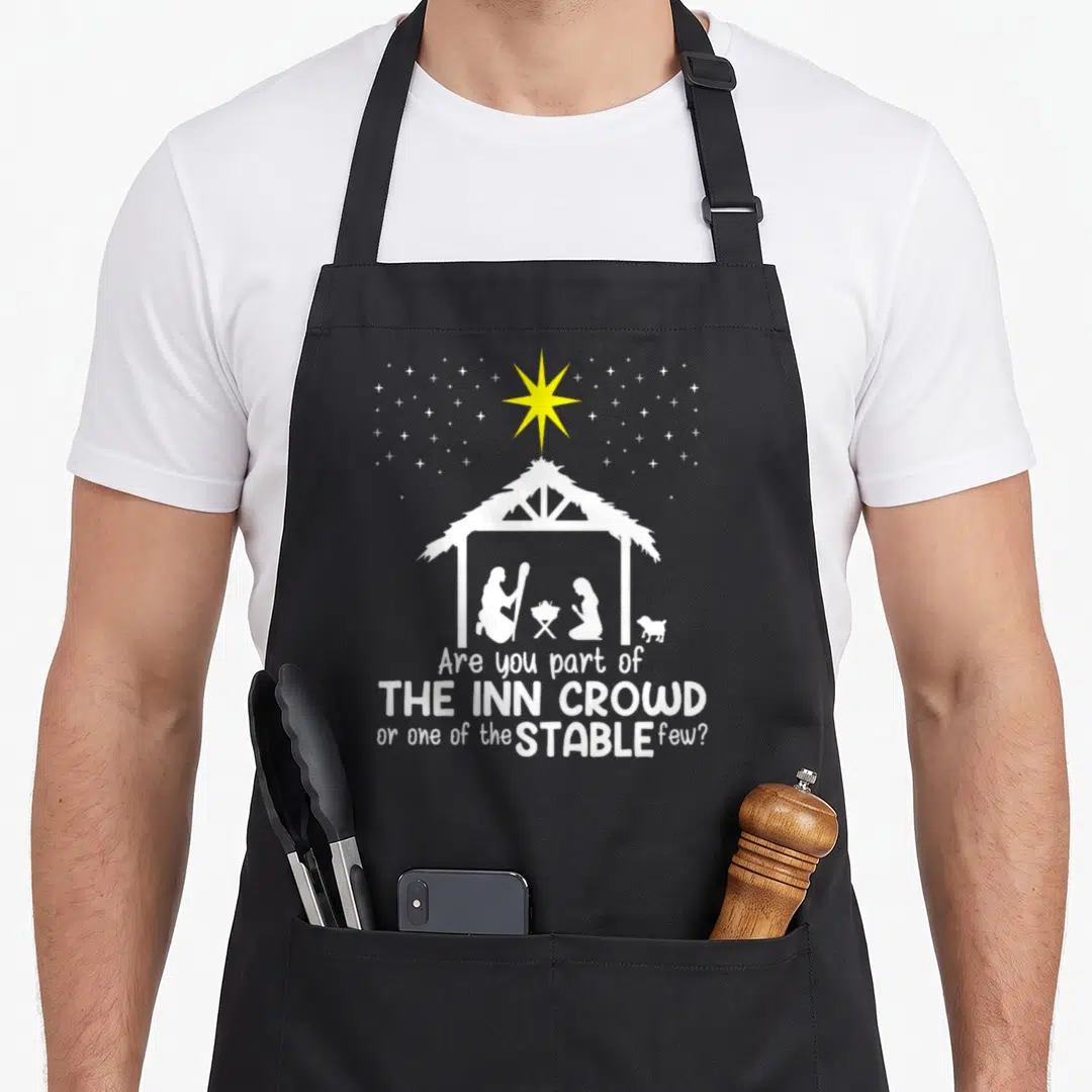 Part womens christmas apron - Nativity stable silhouette with yellow star and inn crowd stable few text — shown on a black cotton canvas apron against a white background