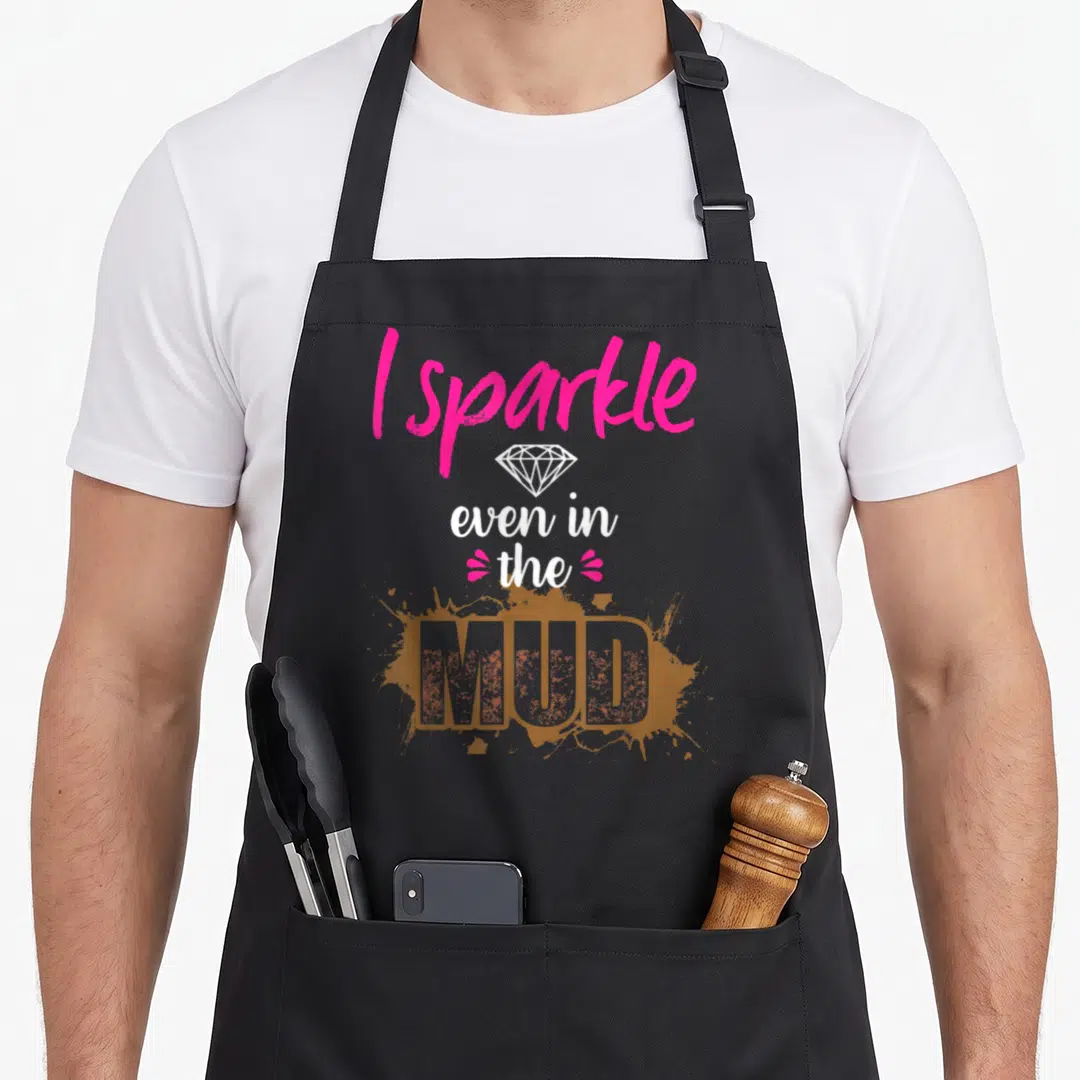 Mud princess apron - Pink and white text reading I sparkle even in the MUD over brown splatter with diamond icon — shown on a black cotton canvas apron against a white background