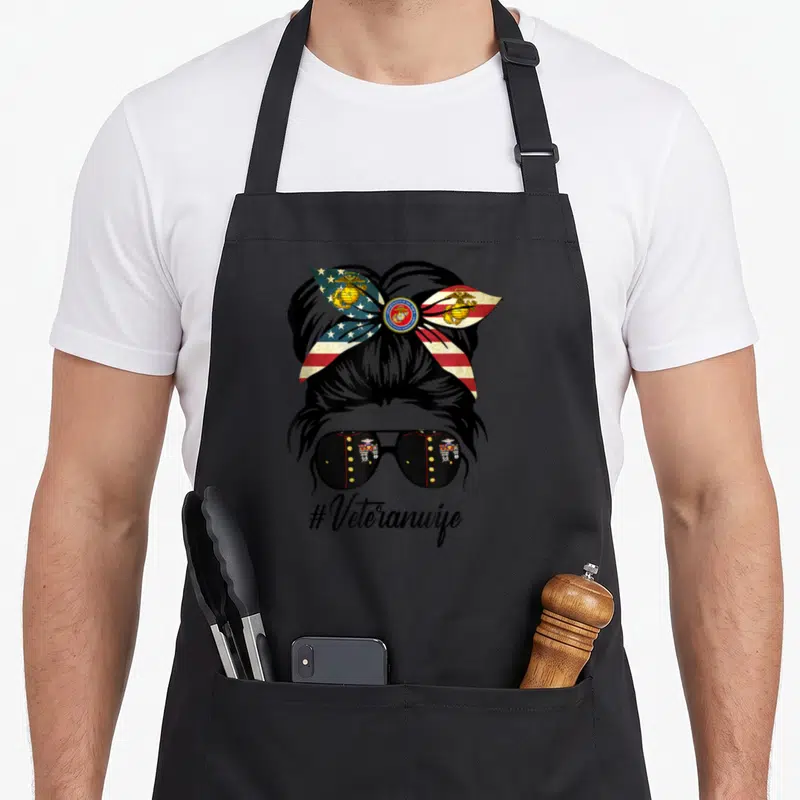 Messy patriotic apron - Stylized woman's head with patriotic bow, uniform sunglasses, and #Veteranwife text — shown on a black cotton canvas apron against a white background