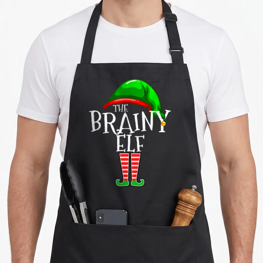 Brainy smart apron - Stacked white text THE BRAINY ELF between green hat and red striped shoes — shown on a black cotton canvas apron against a white background