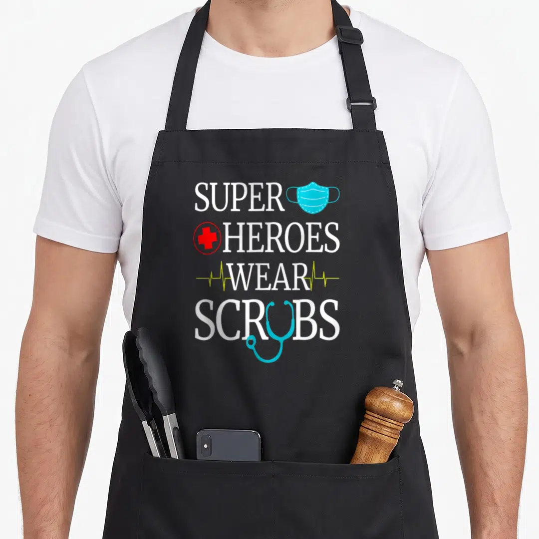 Heroe nursing aprons - Stacked text SUPER HEROES WEAR SCRUBS with mask, cross, heartbeat lines, and stethoscope — shown on a black cotton canvas apron against a white background