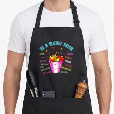 Bucket teacher apron - Smiling pink bucket filled with yellow stars and red hearts surrounded by virtues like love — shown on a black cotton canvas apron against a white background