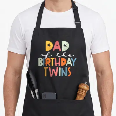 Twin daddy apron - Stacked colorful text reading DAD of the BIRTHDAY TWINS with stars inside letters — shown on a black cotton canvas apron against a white background
