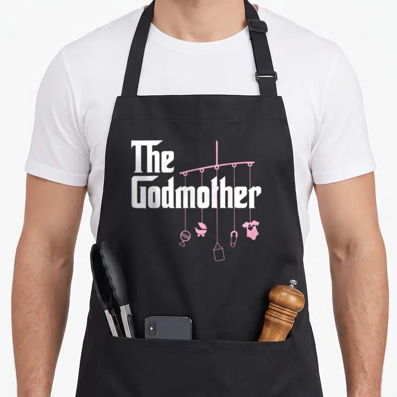 Baby godmother apron - White The Godmother text next to pink mobile with lollipop, carriage, bib, pacifier, and onesie — shown on a black cotton canvas apron against a white background