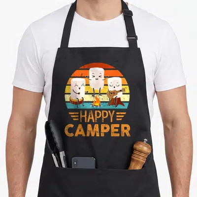 Marshmallow happy camper apron - Three cartoon marshmallows playing music by a campfire under a striped sun with orange — shown on a black cotton canvas apron against a white background