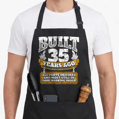35th age birthday apron - White distressed text built 35 years ago on orange ribbon and plaque — shown on a black cotton canvas apron against a white background
