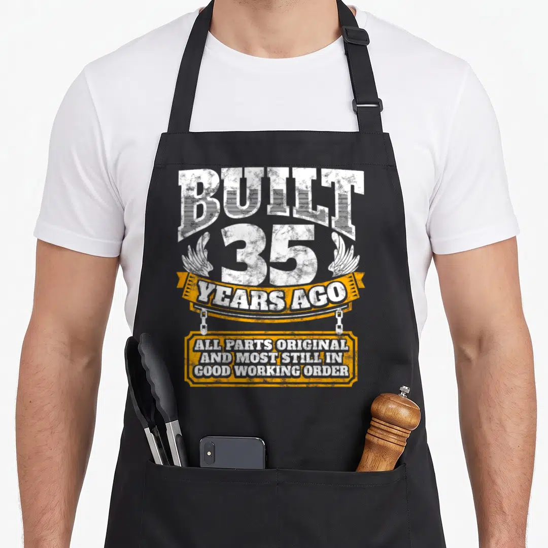35th age birthday apron - White distressed text built 35 years ago on orange ribbon and plaque — shown on a black cotton canvas apron against a white background