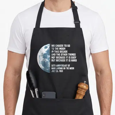 President apollo apron - Half moon graphic beside We Choose to Go to the Moon quote and 1969 anniversary date — shown on a black cotton canvas apron against a white background
