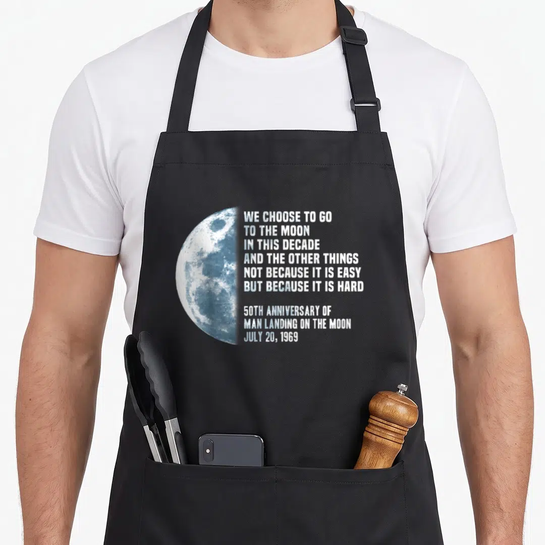 President apollo apron - Half moon graphic beside We Choose to Go to the Moon quote and 1969 anniversary date — shown on a black cotton canvas apron against a white background