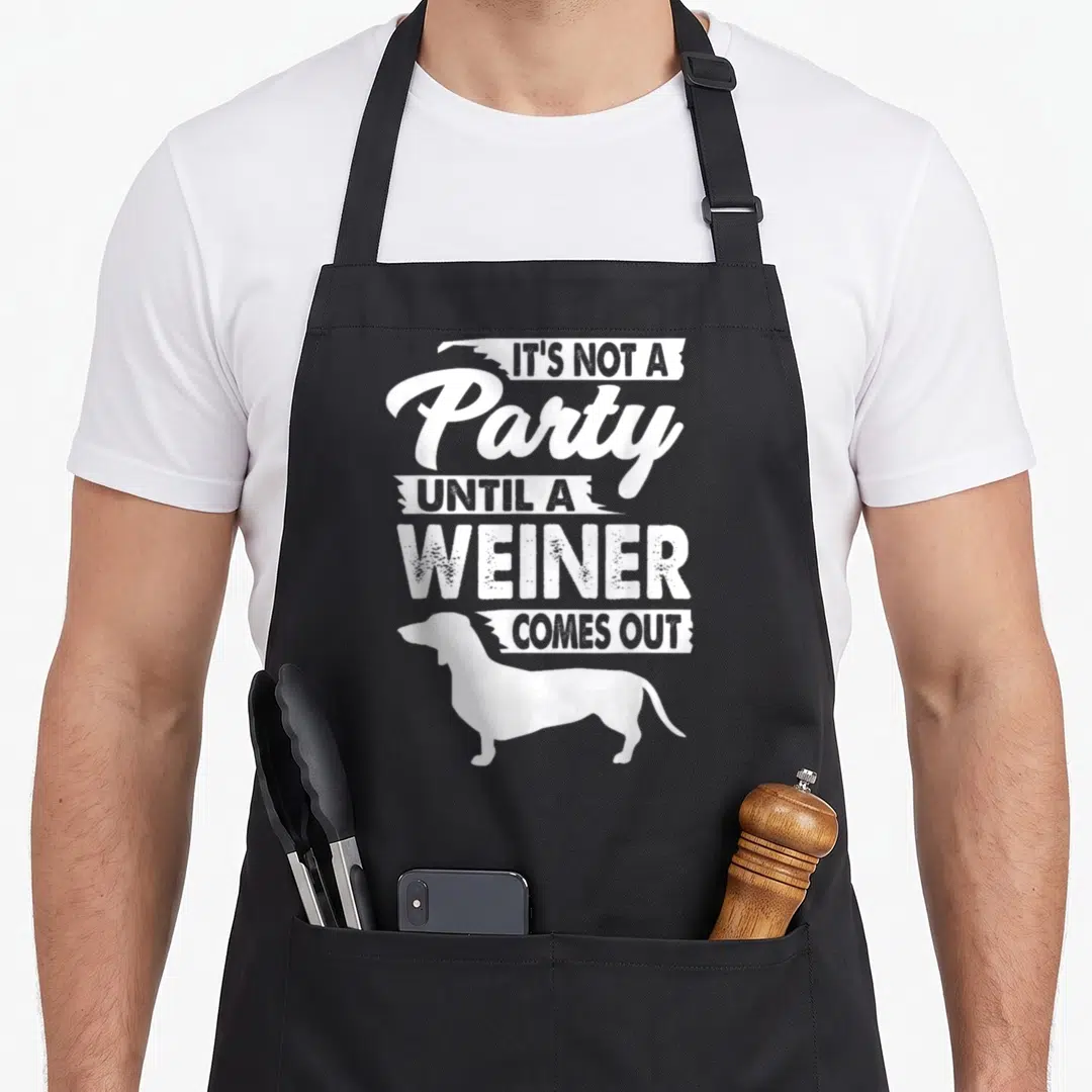 Immature mens joke aprons - White stacked text IT'S NOT A PARTY UNTIL A WEINER COMES OUT above dachshund silhouette — shown on a black cotton canvas apron against a white background