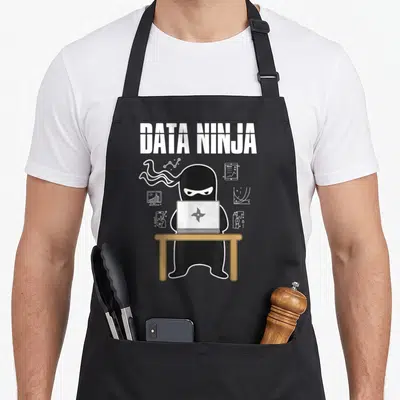 Ninja data analysis analyst apron - Cartoon ninja at brown table with laptop and floating data charts under DATA NINJA — shown on a black cotton canvas apron against a white background