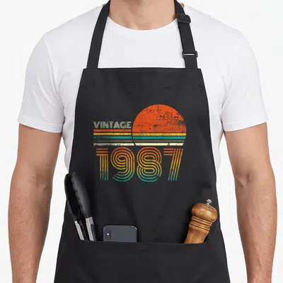 34th quarantine birthday apron - Retro sun graphic with VINTAGE 1987 text in orange yellow teal white stripes — shown on a black cotton canvas apron against a white background