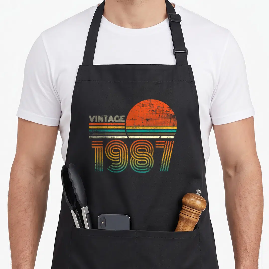 34th quarantine birthday apron - Retro sun graphic with VINTAGE 1987 text in orange yellow teal white stripes — shown on a black cotton canvas apron against a white background