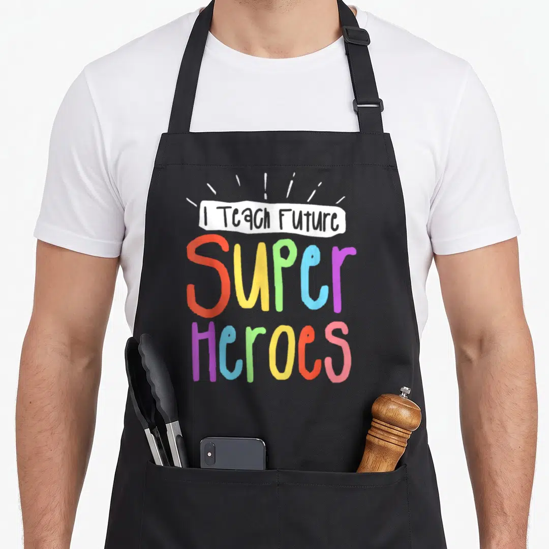 Teach future teacher apron - I Teach Future Super Heroes text in rainbow colors with white banner and rays — shown on a black cotton canvas apron against a white background