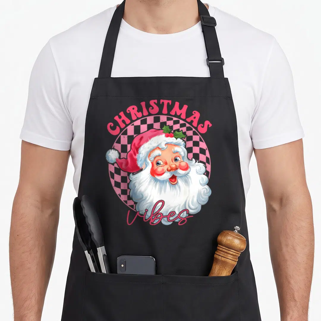 Groovy santa apron - Cartoon Santa head on pink checkerboard with CHRISTMAS and Vibes text in red — shown on a black cotton canvas apron against a white background