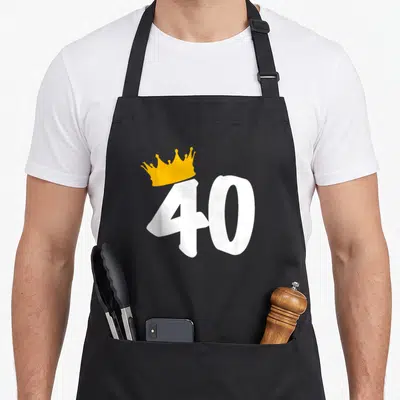 40th queen of baking apron - White number 40 with tilted yellow crown on top of the digit four — shown on a black cotton canvas apron against a white background