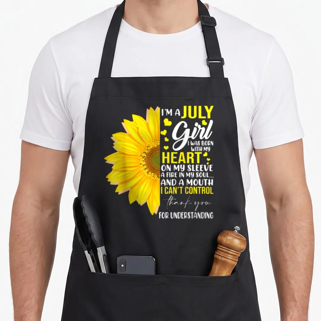 July sunflower apron - Half sunflower next to stacked text reading I'm a July Girl with small hearts — shown on a black cotton canvas apron against a white background