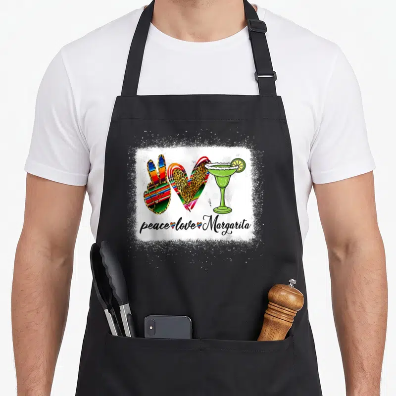 Peace margarita apron - Peace sign, leopard heart, and green margarita glass above script text on white splatter — shown on a black cotton canvas apron against a white background