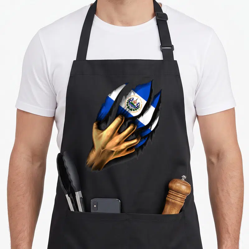 Cute salvador salvadoran apron - Gold hand tearing through surface revealing Salvadoran flag and national text — shown on a black cotton canvas apron against a white background