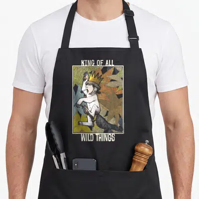 Wild king apron - Crowned monster in white frame with yellow orange grey leaves and KING OF ALL WILD THINGS text — shown on a black cotton canvas apron against a white background