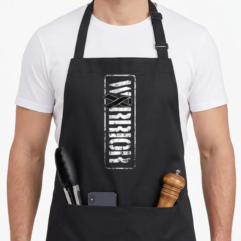 Melanoma military apron - Vertical WARRIOR text in white distressed stencil inside a rectangle with a black awareness — shown on a black cotton canvas apron against a white background