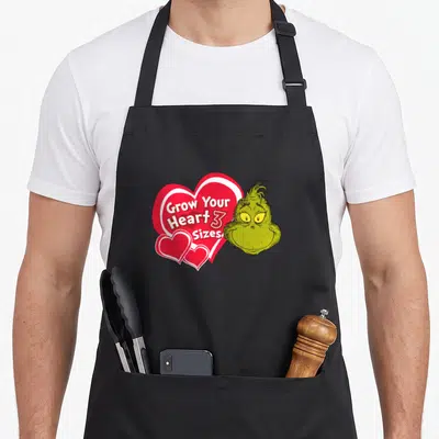 Grow holiday apron - The Grinch head next to red heart with Grow Your Heart 3 Sizes text — shown on a black cotton canvas apron against a white background