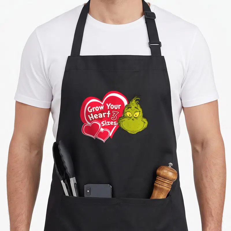Grow holiday apron - The Grinch head next to red heart with Grow Your Heart 3 Sizes text — shown on a black cotton canvas apron against a white background
