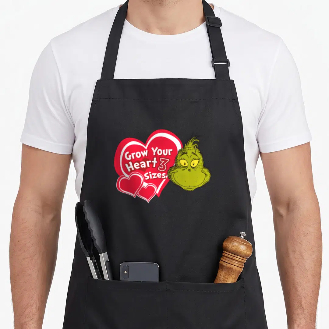 Grow holiday apron - The Grinch head next to red heart with Grow Your Heart 3 Sizes text — shown on a black cotton canvas apron against a white background