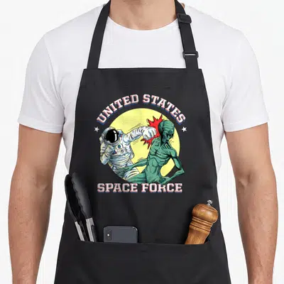 State funny alien apron - Retro astronaut punching green alien with UNITED STATES SPACE FORCE text over yellow circle — shown on a black cotton canvas apron against a white background