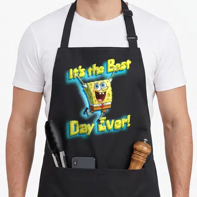 Squarepant general apron - SpongeBob jumping between arched yellow text It's the Best Day Ever on blue halftone — shown on a black cotton canvas apron against a white background